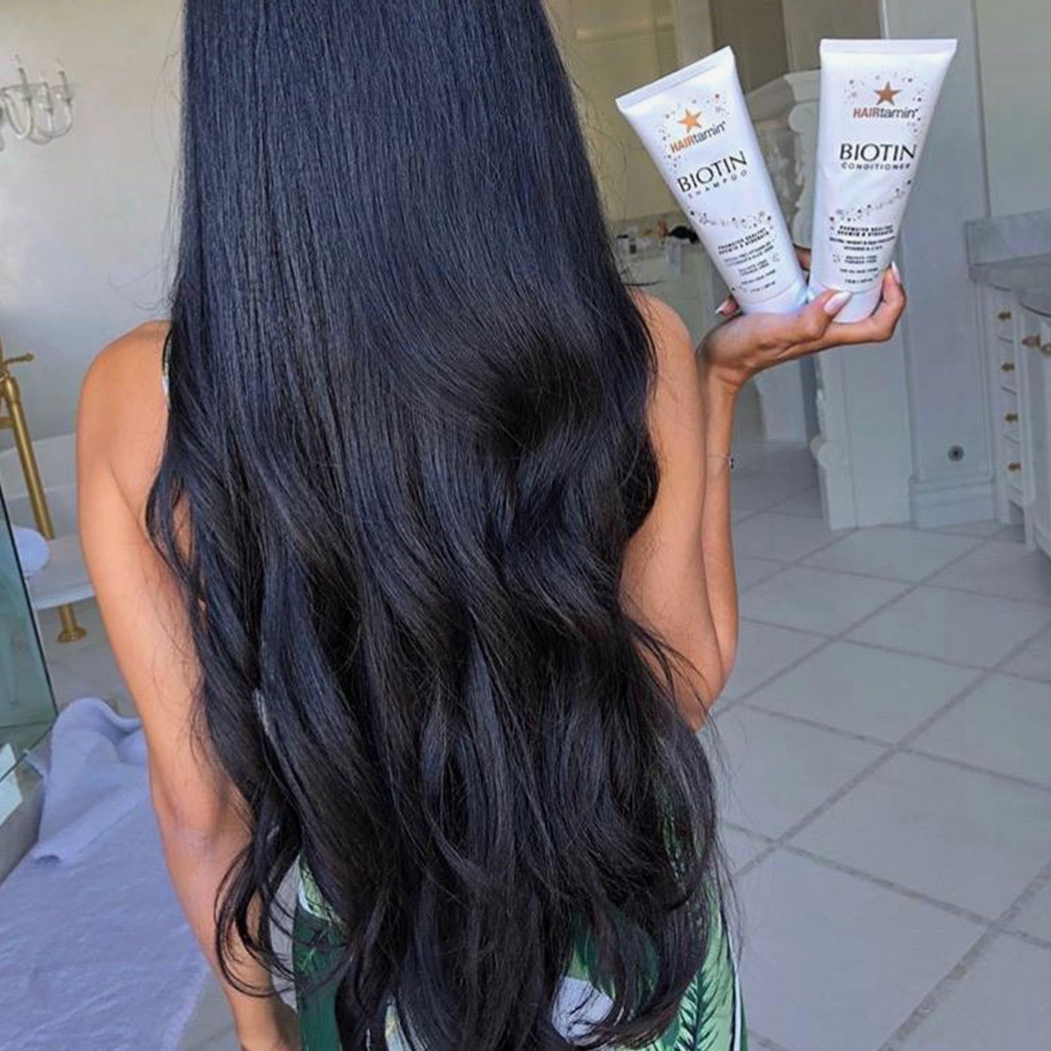 Zwei weiße Tuben "HAIRtamin Biotin Shampoo" in Händen. Schwarzes, glattes Haar im Vordergrund.