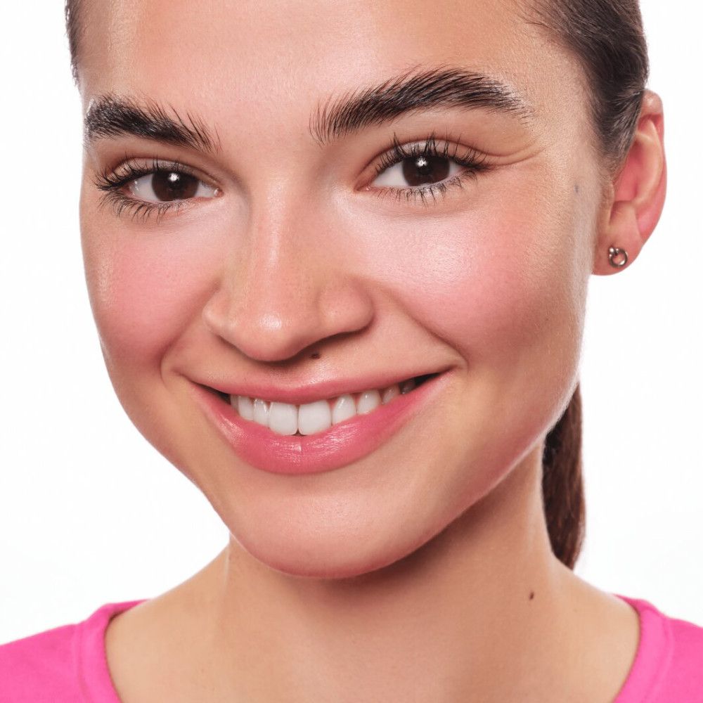 Porträt einer lächelnden Frau mit gebräunter Haut. Sie trägt rosa Oberteil. Deutliche Augenbrauen und Lippenstift.