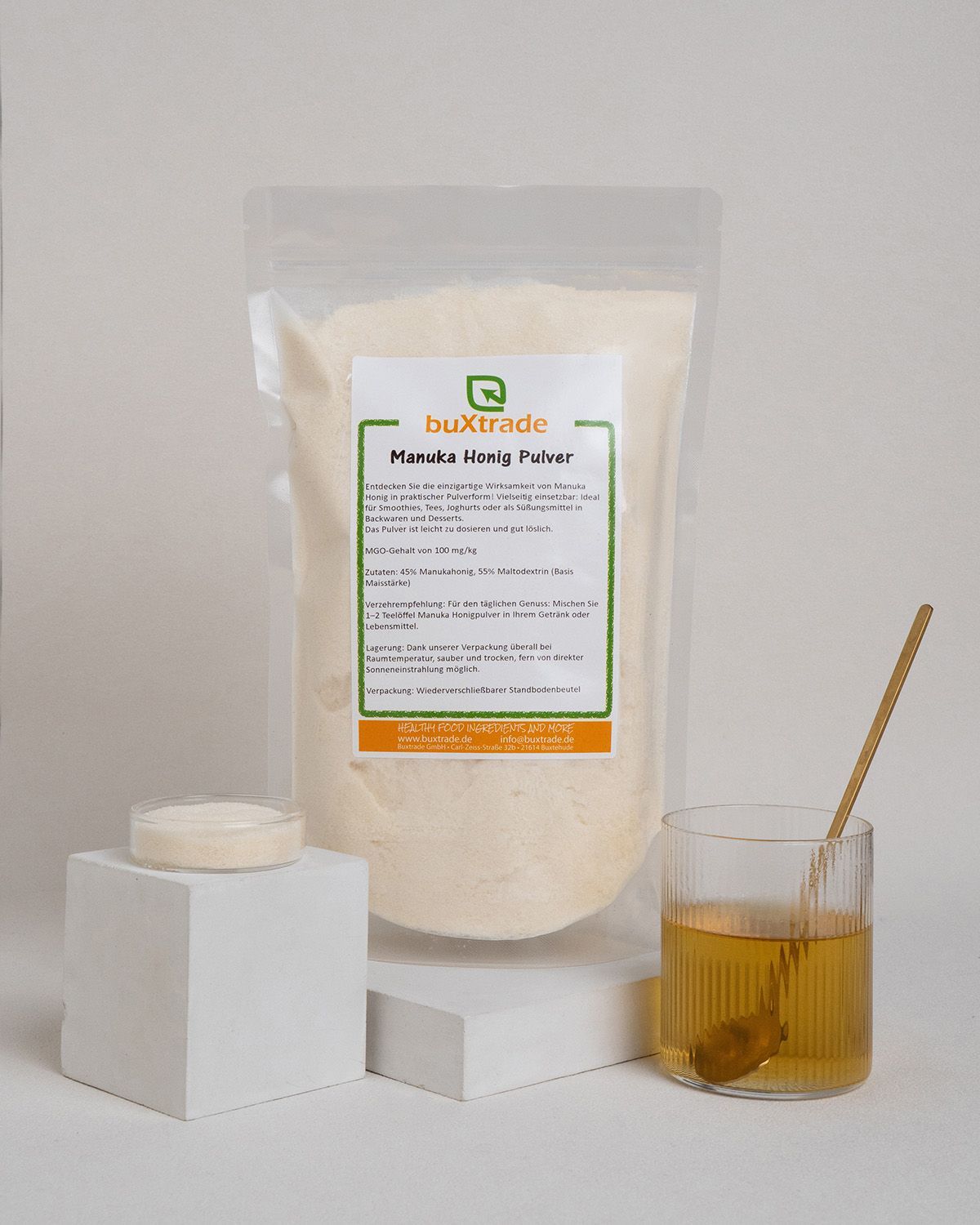 Beutel mit Pulver, Glas mit Flüssigkeit, Löffel. Aufdruck: buXtrade Manuka Honig Pulver. Weißes Podest.