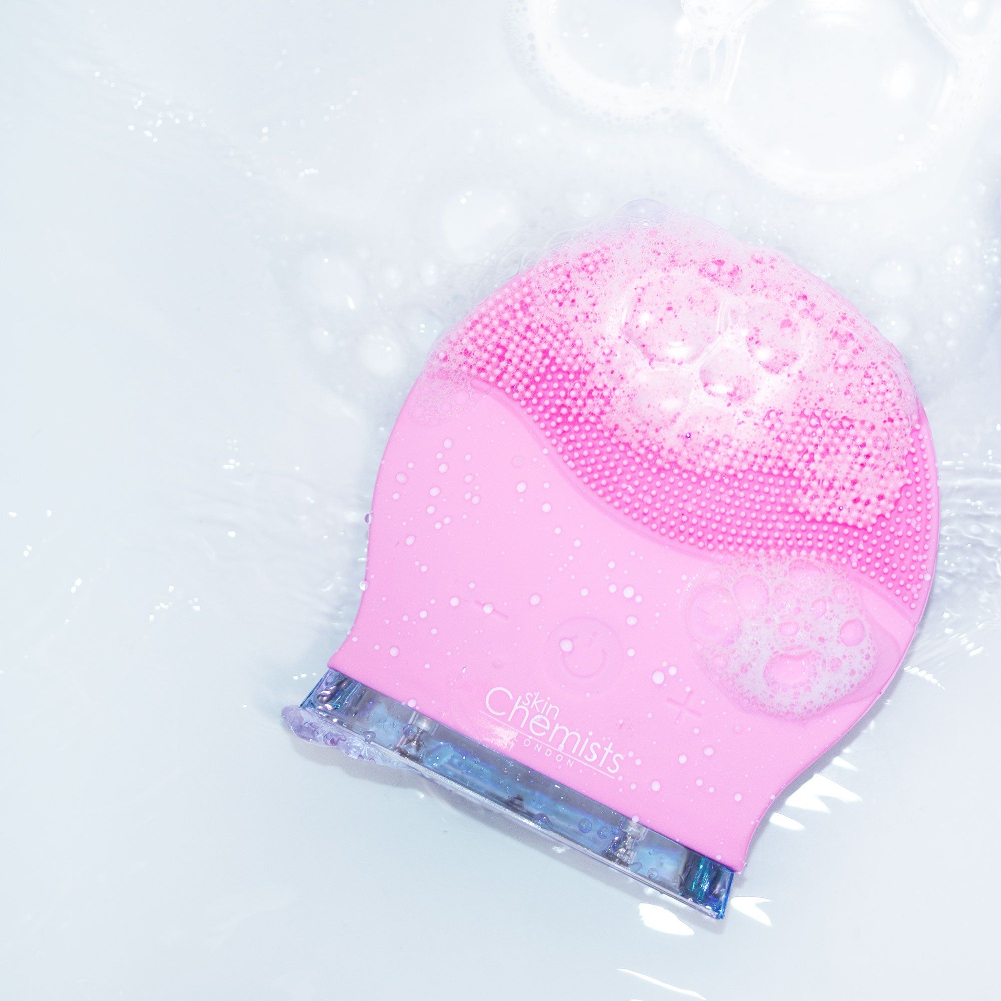 Rosa Gesichtsreinigungsbürste in Wasser mit Seifenblasen. Bürstenkopf mit Silikonborsten. Produktname: SkinChemists.