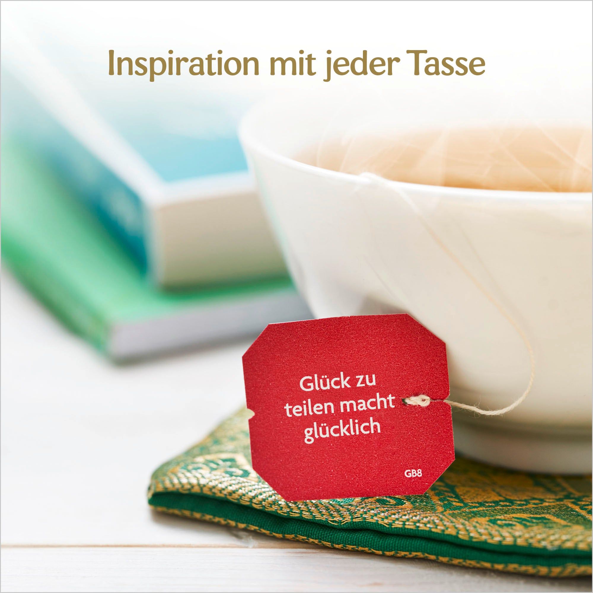 Weiße Tasse mit Teebeutel. Rotes Etikett: Glück zu teilen macht glücklich. Bücher im Hintergrund.
