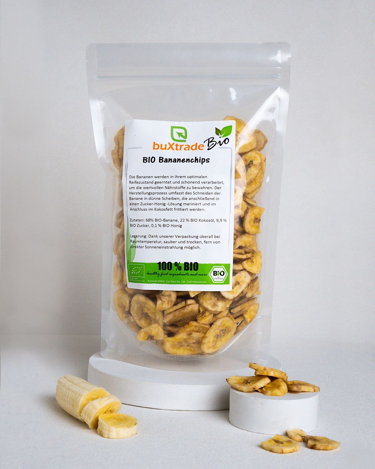 Klarsichtbeutel mit Bio Bananenchips. Aufkleber mit Produktinformationen. Daneben geschnittene Banane und Chips auf weißem Podest.