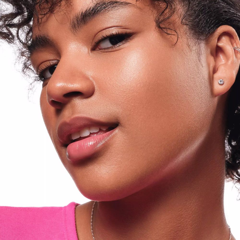 Nahaufnahme einer Frau mit gehighlighteten Wangenknochen. Sie trägt ein rosa Oberteil. Ihr Haar ist lockig.