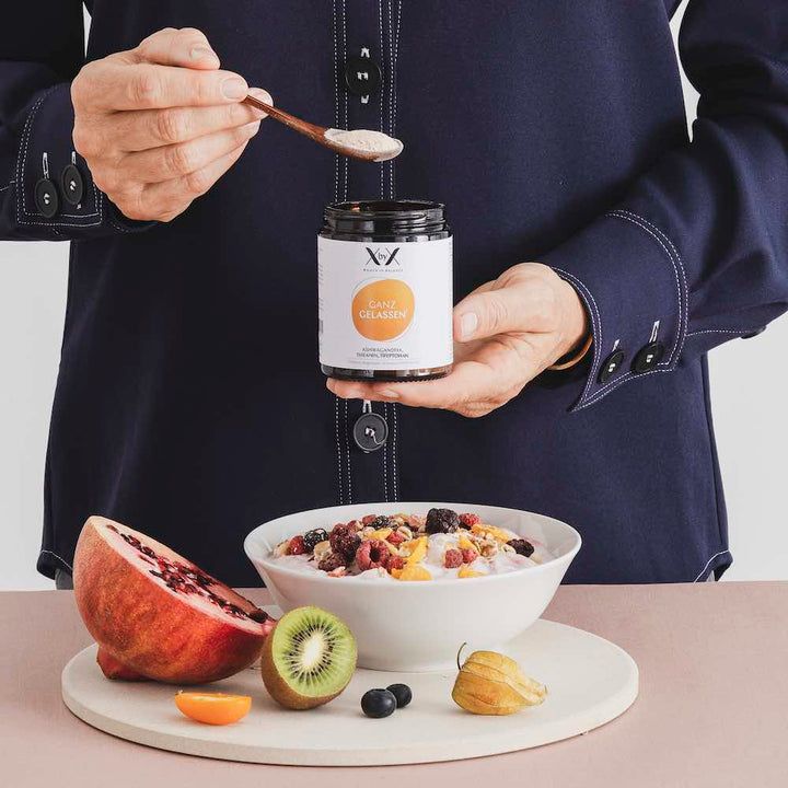 Person hält Löffel mit Pulver über einer Schüssel mit Obst und dem Produkt Ganz Gelassen.