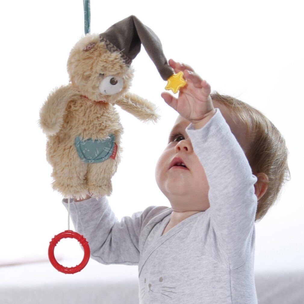 Ein Baby blickt auf eine beige Bär-Spieluhr mit Mütze und Stern. Ein roter Ring hängt an der Spieluhr.