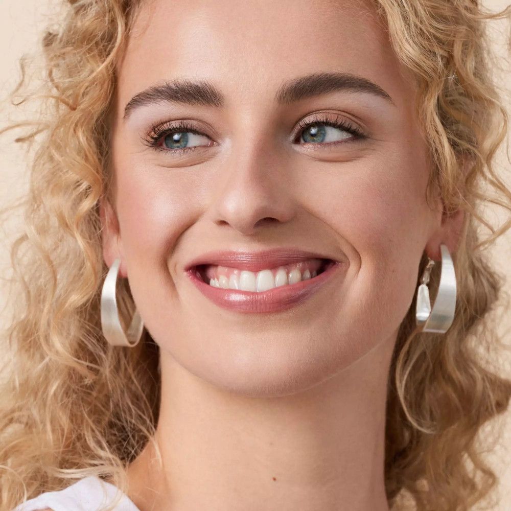 Porträt einer lächelnden Frau mit blonden Locken. Sie trägt silberne Ohrringe und Make-up.