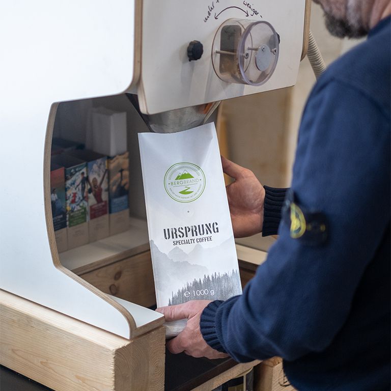 Kaffeebohnen-Verpackung. Weiß, mit Logo und Text. Person hält die Packung. 1000g.