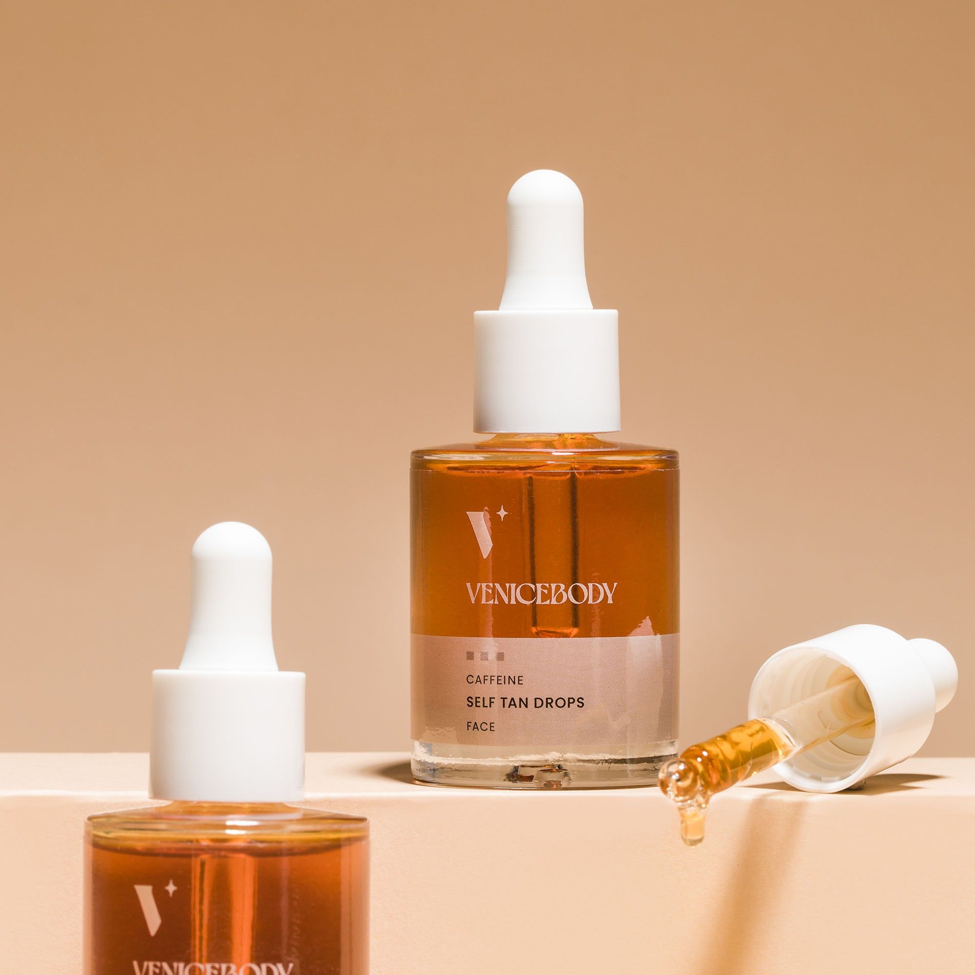 Mehrere Flaschen mit brauner Flüssigkeit und weißen Pipetten. Eine Pipette liegt daneben. Aufschrift: VENICEBODY, Caffeine Self Tan Drops Face.