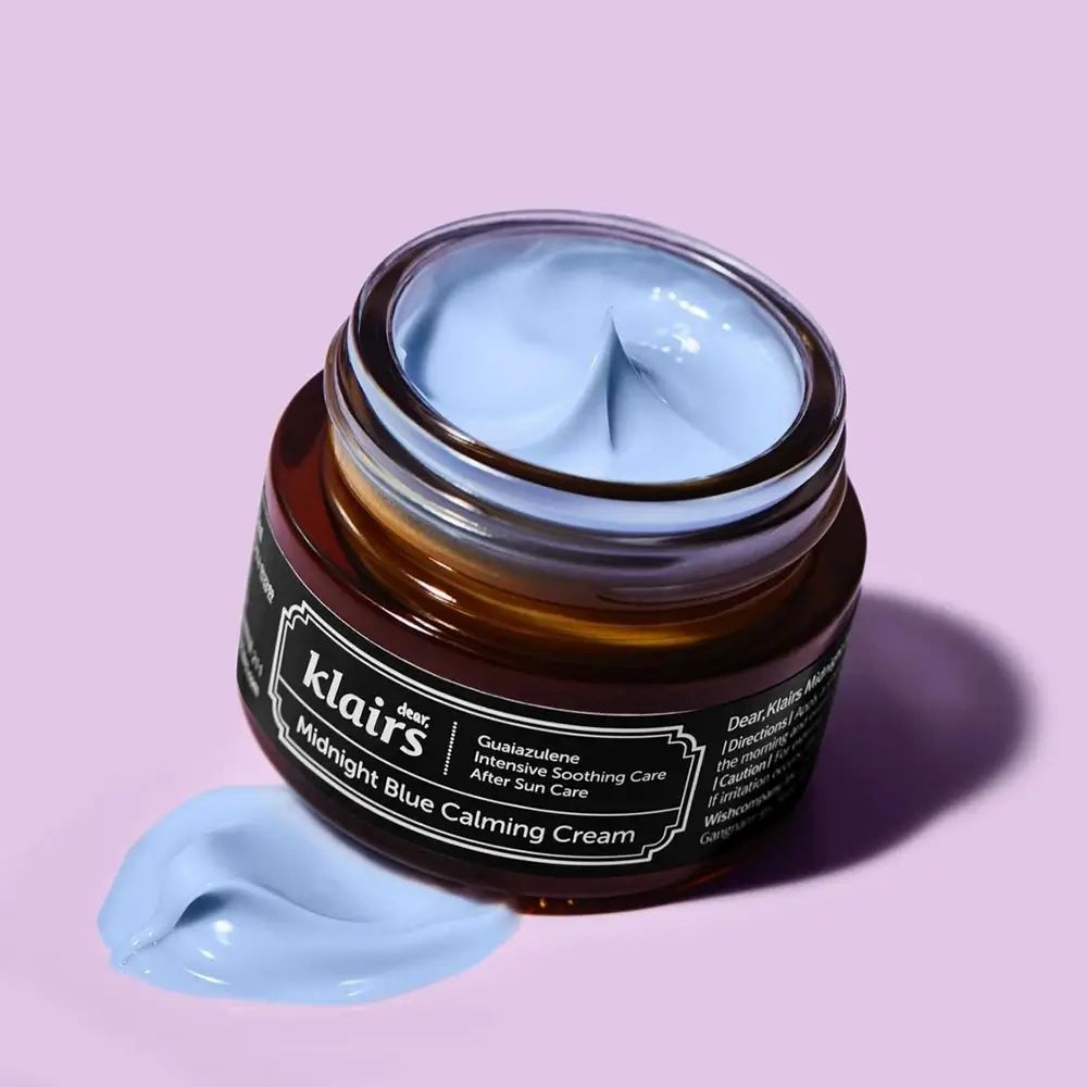 Geöffnetes Creme-Glas, blaue Creme sichtbar. Produktname und Markenname auf dem Etikett. Creme-Klecks daneben.