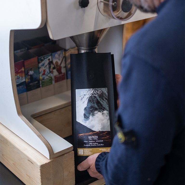 Person hält eine schwarze Kaffeepackung. Text: BERGBRAND, Ursprung, Filter, 250g. Zutaten: Kakao, Haselnuss, Schokolade, 100% Arabica.