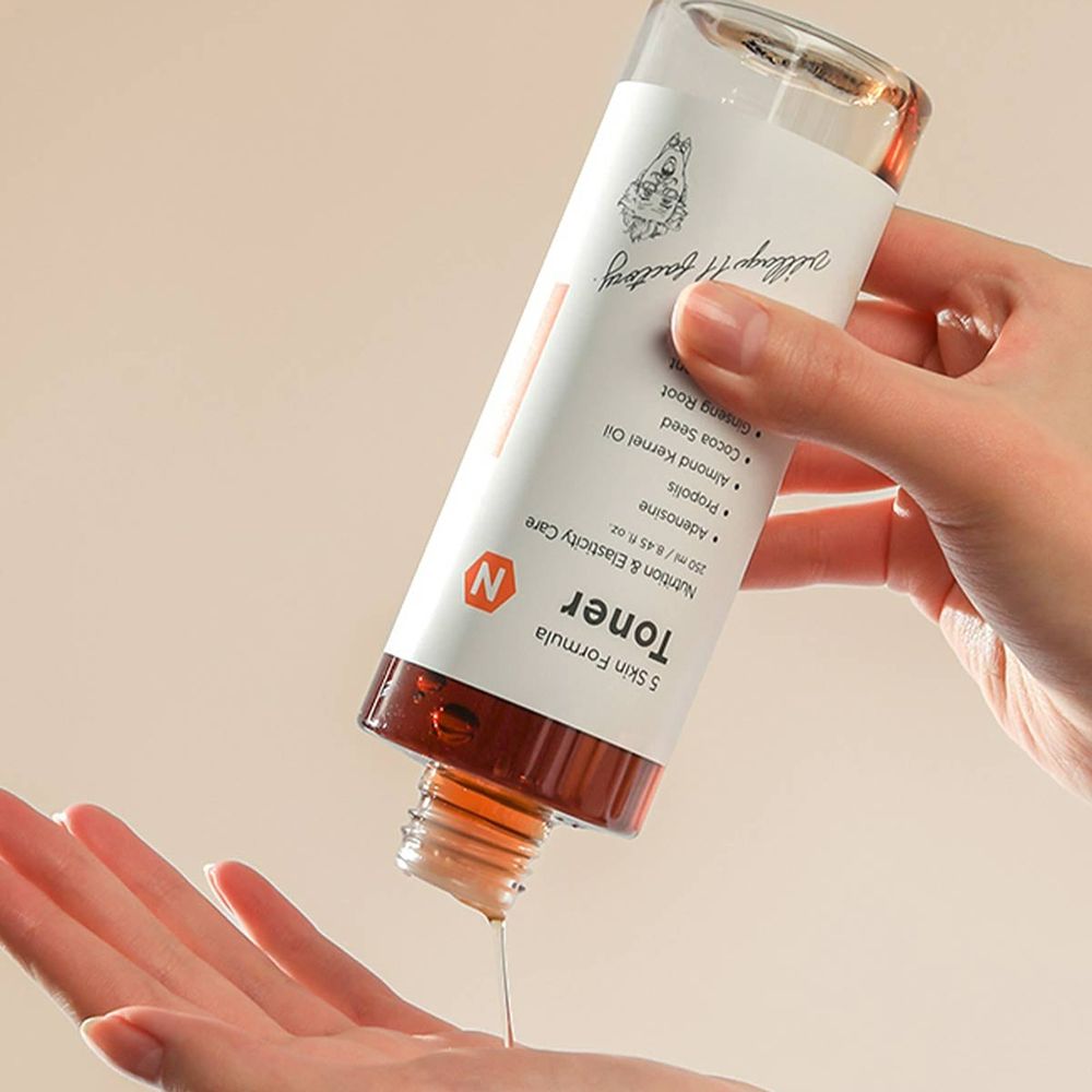 Flasche wird über eine Hand gehalten. Flüssigkeit tropft heraus. Etikett mit Text: Toner, 5 Skin Formula, N. Orangefarbener Streifen.