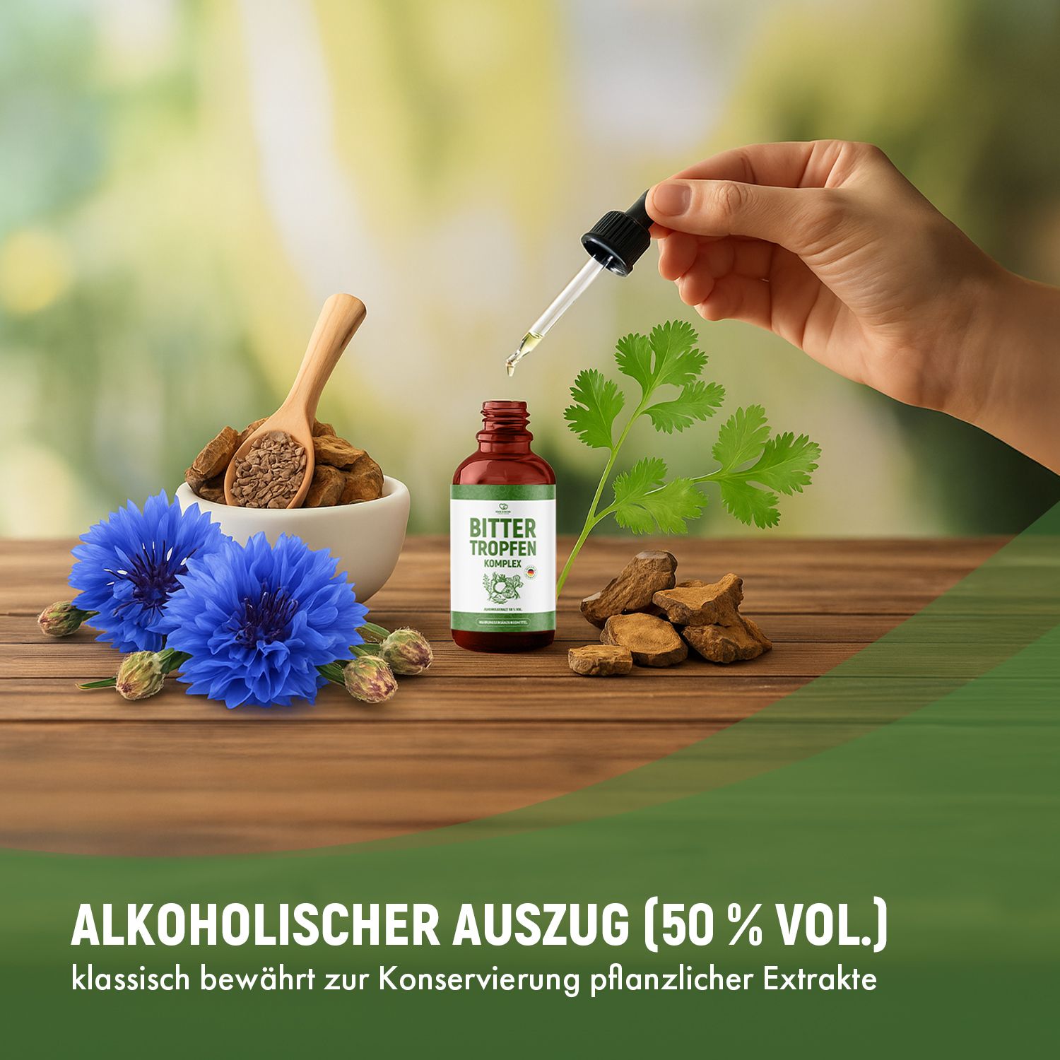 Hand hält Tropfer über einer Flasche Bitter Tropfen. Auf einem Holztisch: Kräuter, blaue Blüten, Samen. Alkoholgehalt 50% Vol.