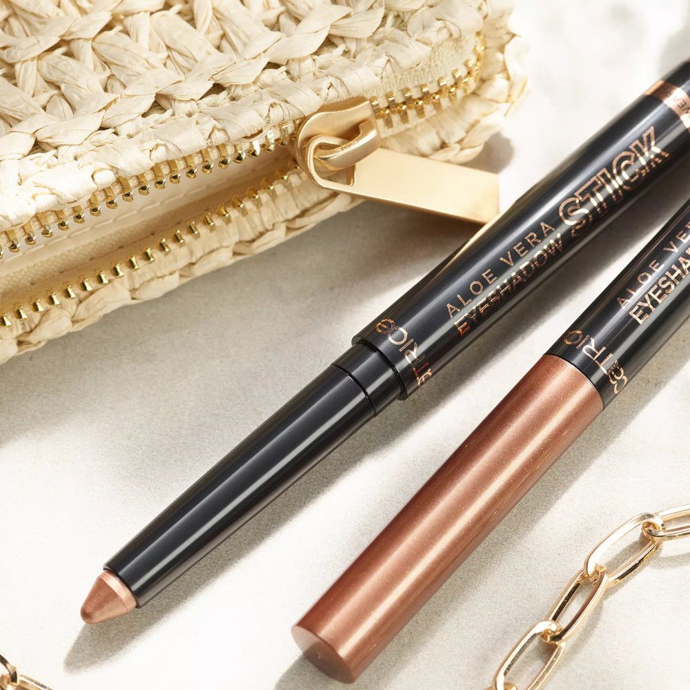 Zwei Catrice Aloe Vera Eyeshadow Sticks neben einer Clutch und einer Kette. Ein Stick ist geöffnet.
