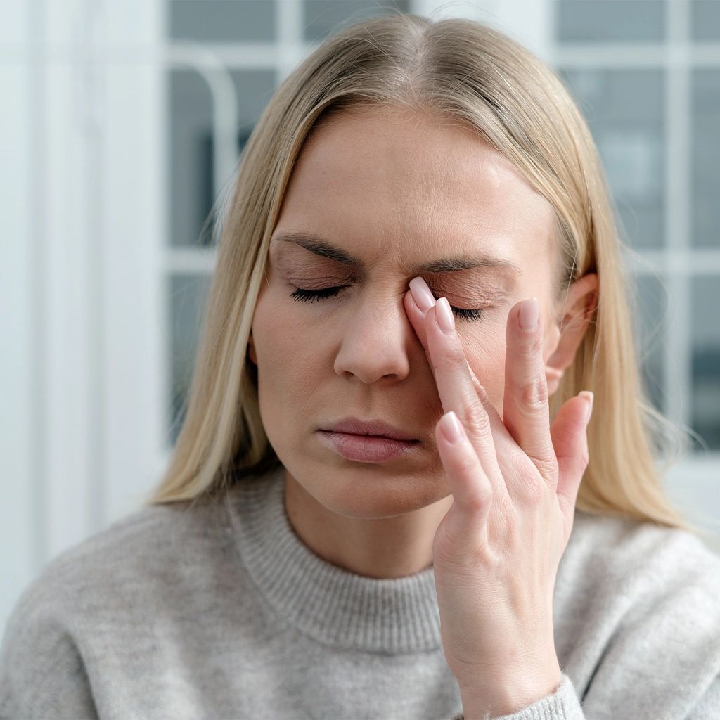 Frau mit geschlossenen Augen, berührt ihr Auge mit der Hand. Helle Kleidung, heller Hintergrund.