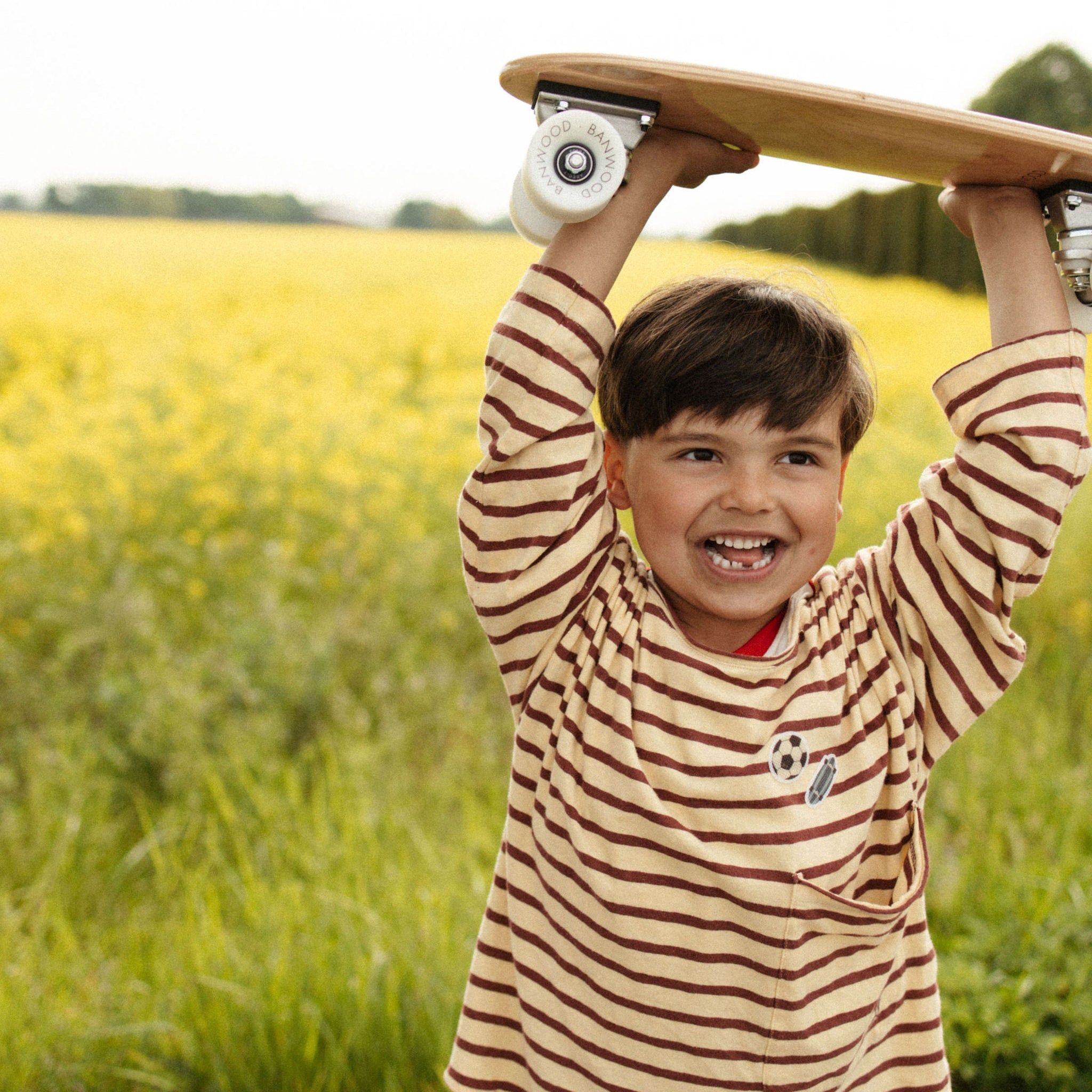 Junge mit Skateboard und gestreiftem Shirt im Freien, gelbes Feld im Hintergrund.
