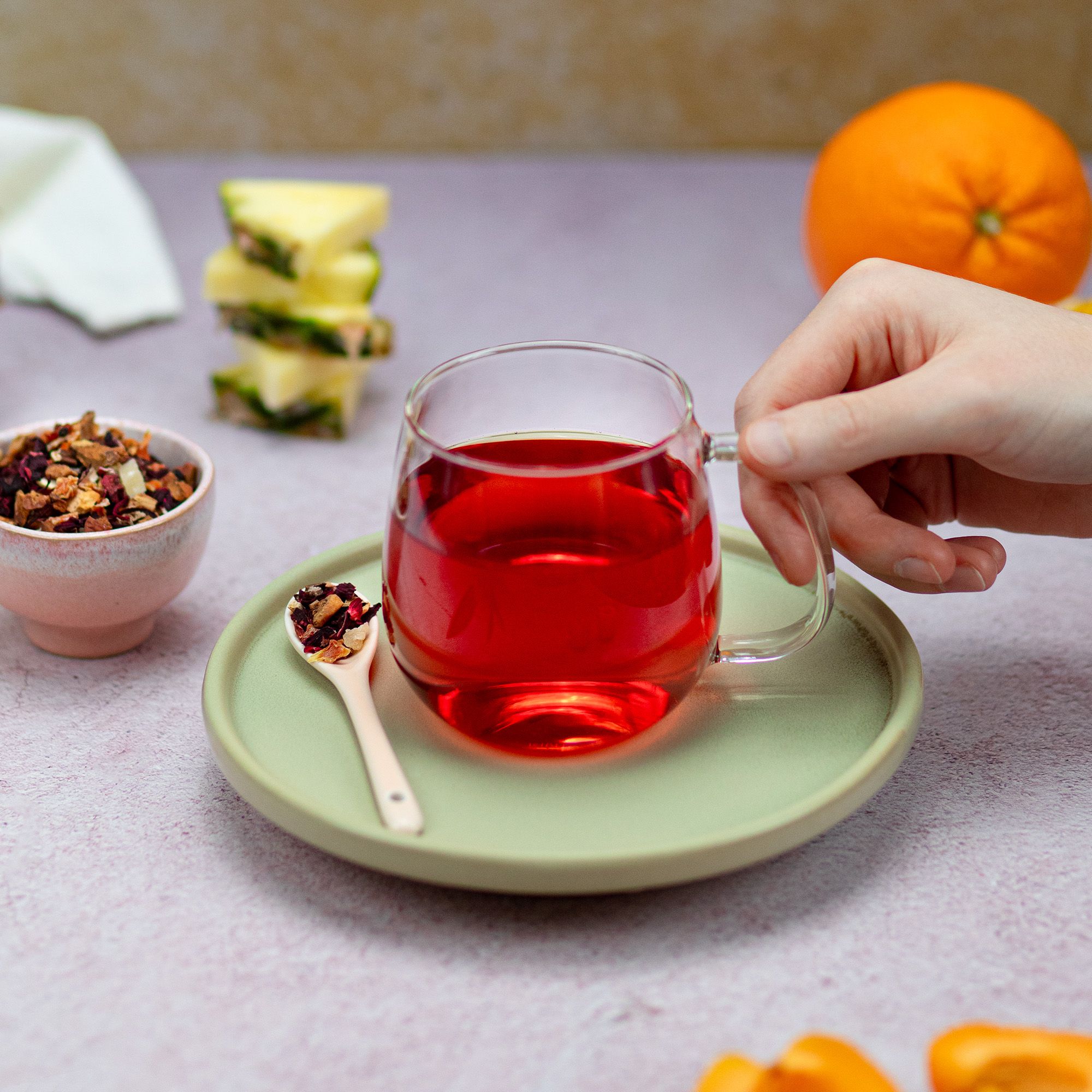 Glasbecher mit rotem Tee, auf Teller mit Löffel. Früchtetee-Mischung in Schale und Obst. Hand hält Becher.