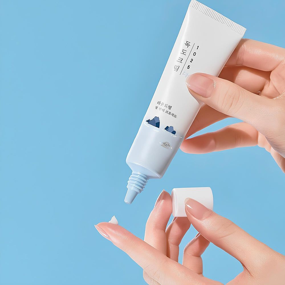 Weiße Tube mit blauer Aufschrift und Illustration. Eine Hand hält die Tube, die andere den Deckel. Creme wird herausgedrückt.