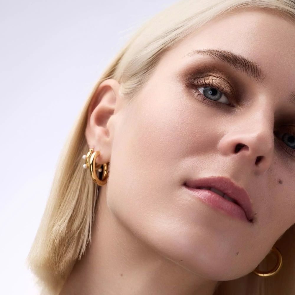 Nahaufnahme einer Frau mit blonden Haaren und goldenen Creolen. Aufgetragenes Augen-Make-up.