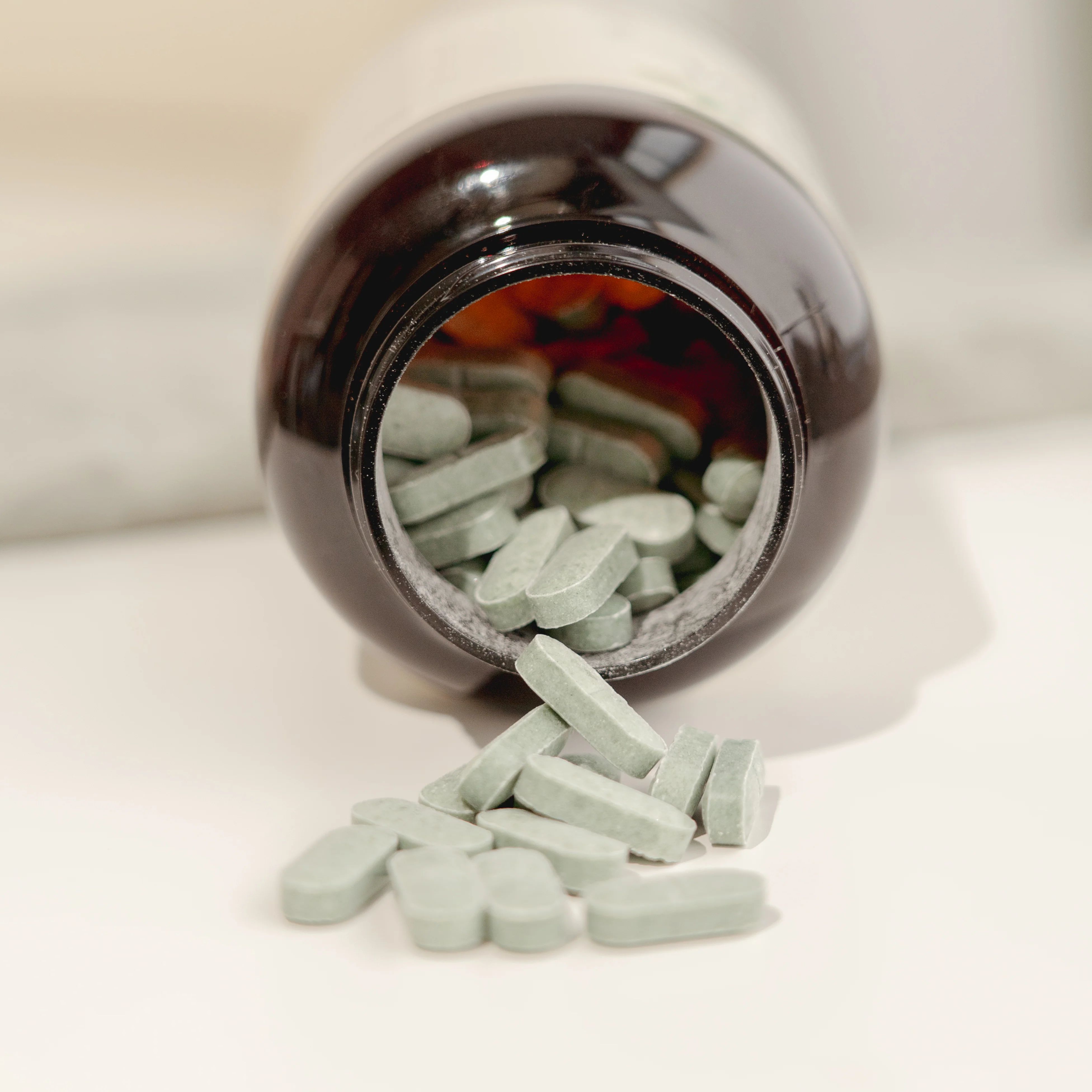 Braune Tablettenflasche, geöffnet, Tabletten fallen heraus. Hellgrüne, längliche Tabletten liegen verstreut.