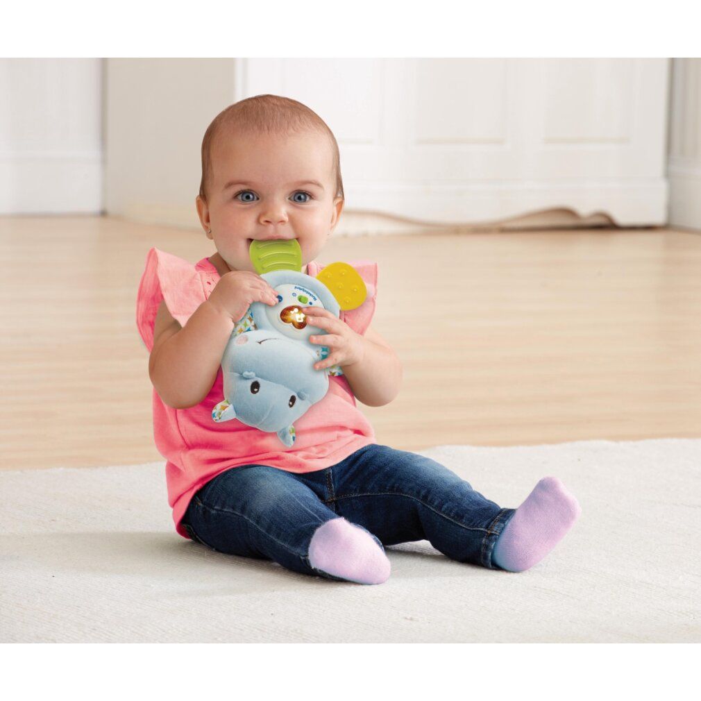 Baby sitzt und hält ein blaues Stoff-Nilpferd-Spielzeug im Mund. Das Spielzeug hat bunte Details und einen Knopf.