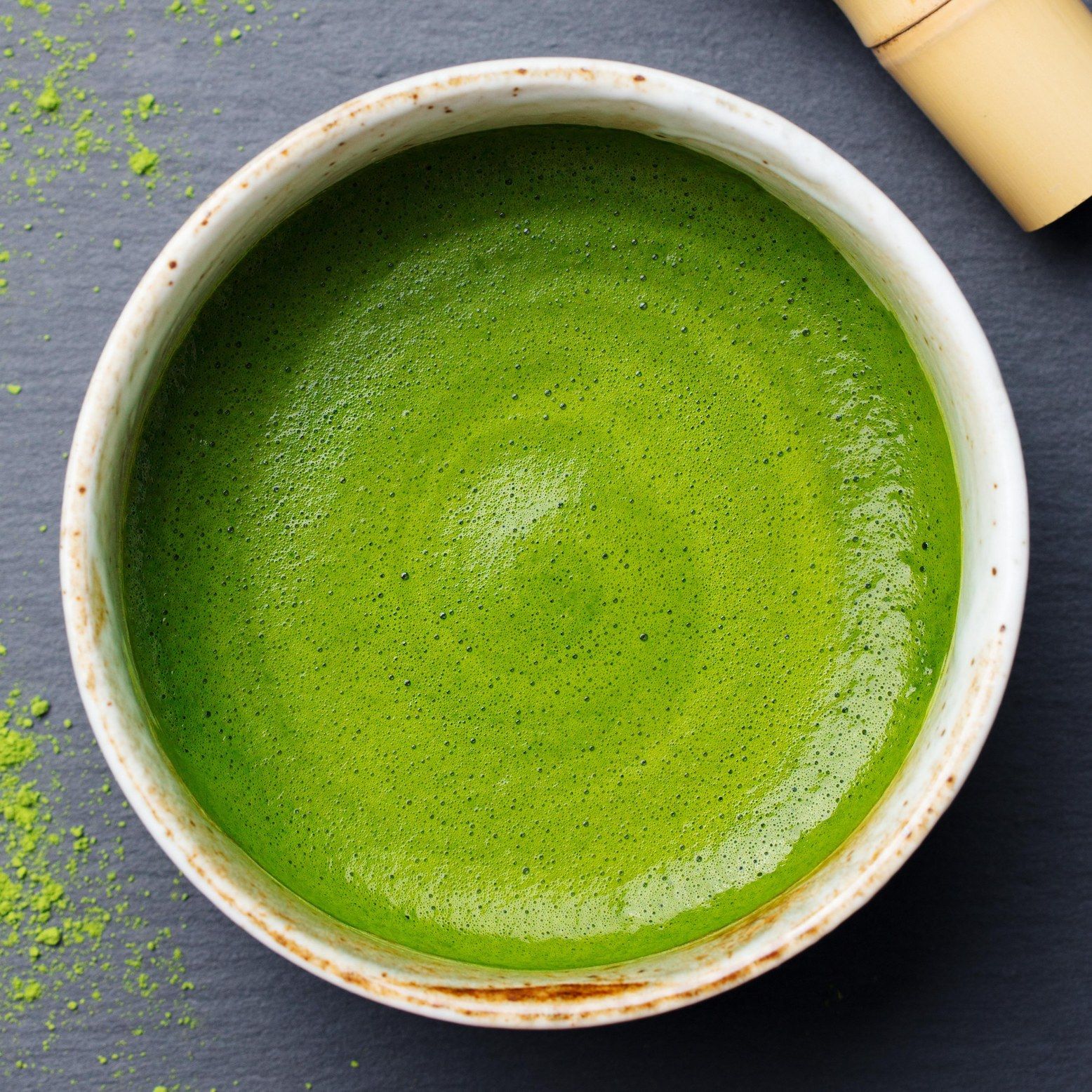 Schale mit Matcha-Tee, cremige Textur, Bambusbesen daneben, auf dunklem Untergrund.