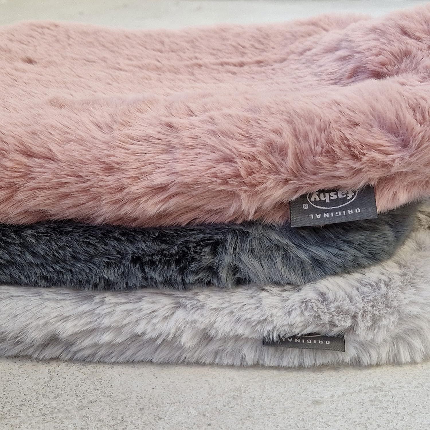 Drei Wärmflaschen mit flauschigen Bezügen. Rosa, grau und hellgrau. Rechteckige Form. Etiketten mit 'Fashy'.
