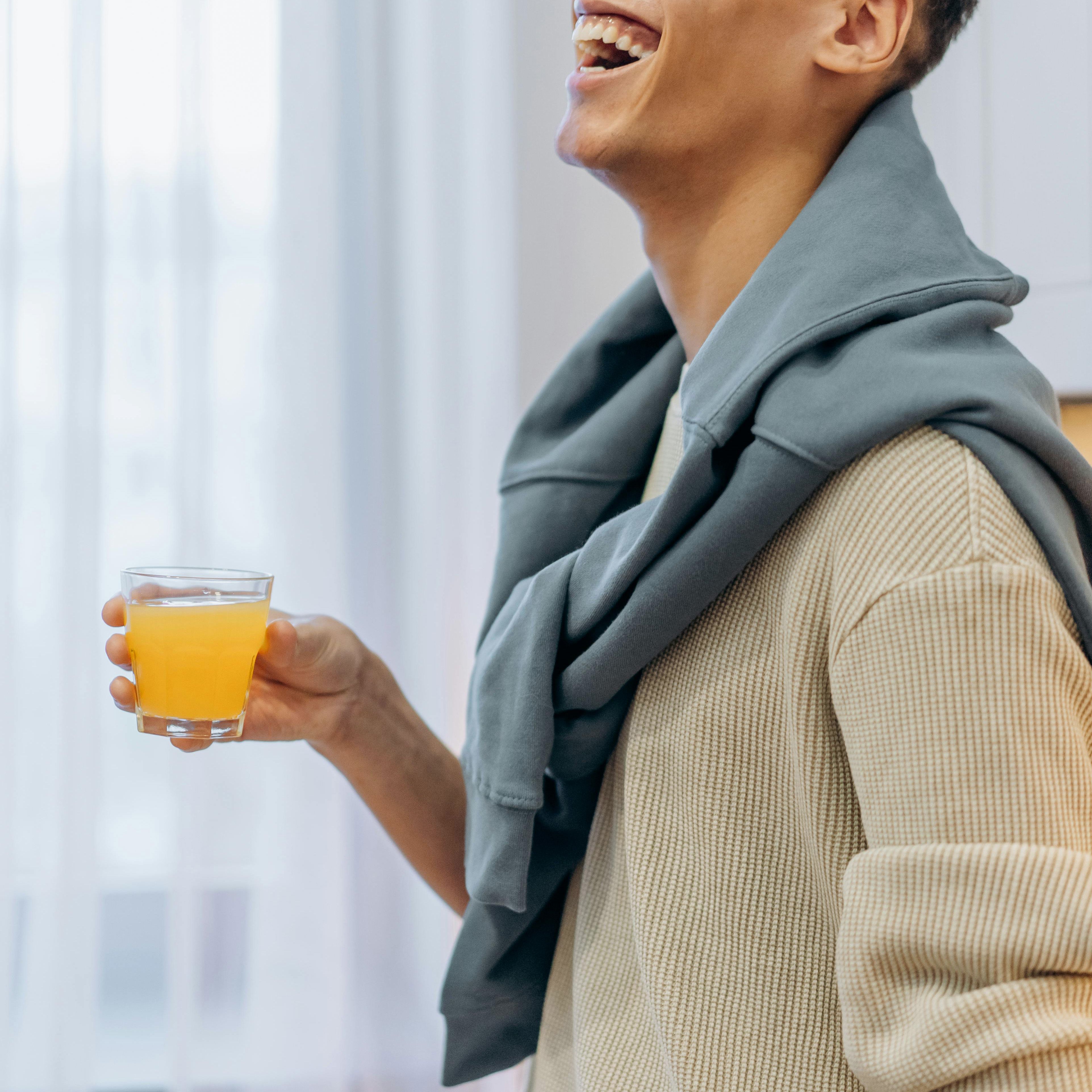 Mann hält Glas mit orangefarbenem Getränk. Er lacht. Er trägt einen beigen Pullover und einen blauen Pullover über den Schultern.
