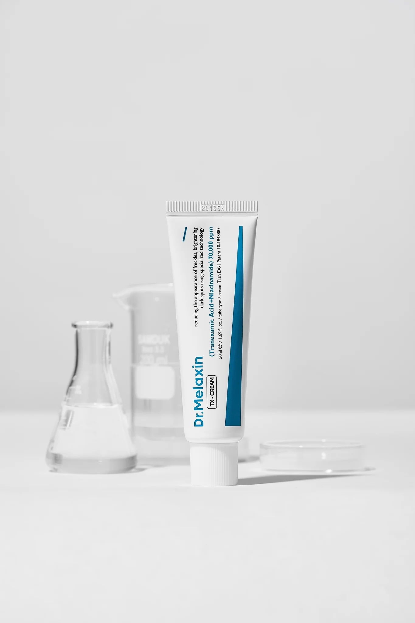 Weiße Tube mit blauer Beschriftung. Aufschrift: Dr.Melaxin TX-CREAM. Enthält Tranexamsäure und Niacinamid.