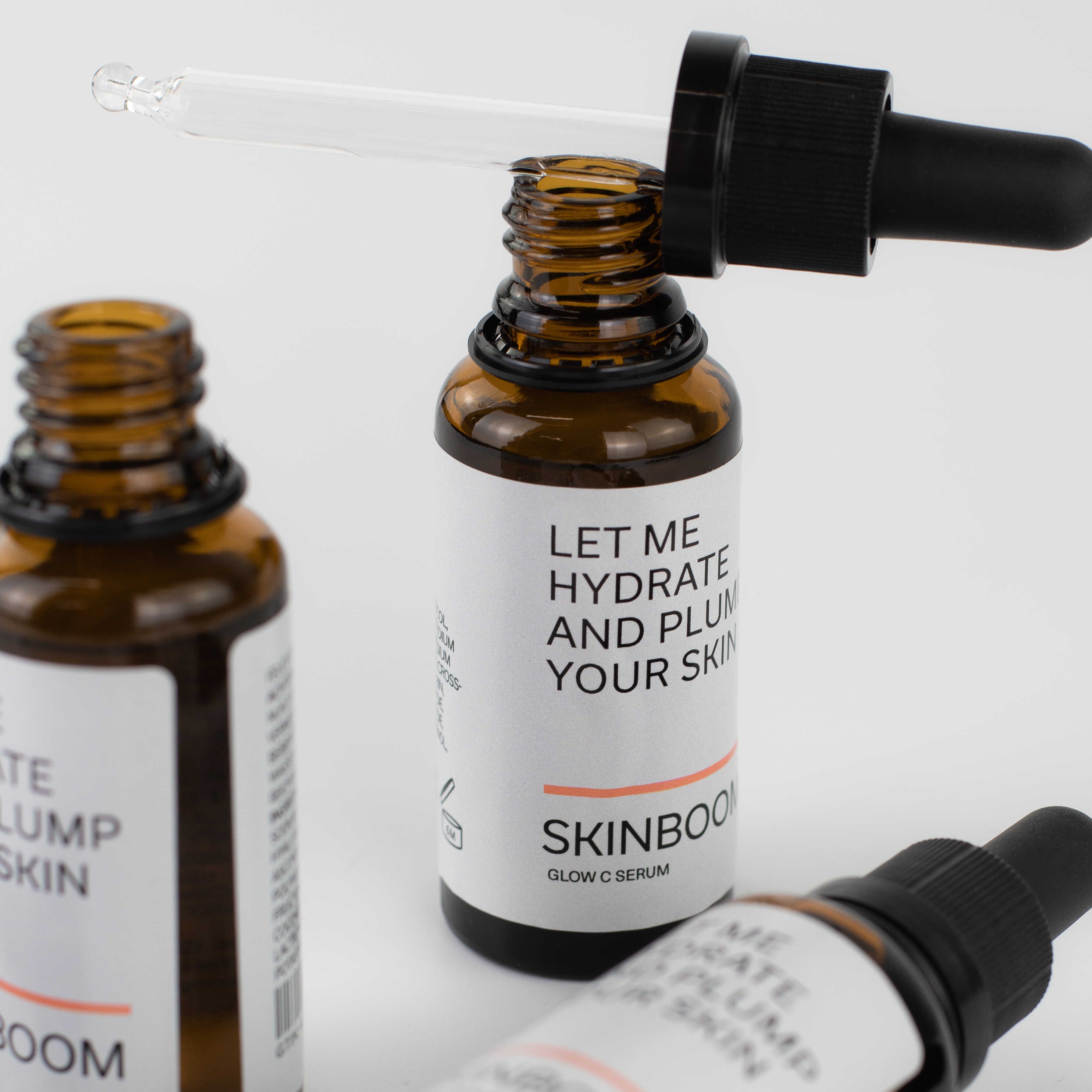 Drei braune Glasflaschen mit Pipetten. Etikett mit Text: LET ME HYDRATE AND PLUMP YOUR SKIN. SKINBOOM Glow C Serum.
