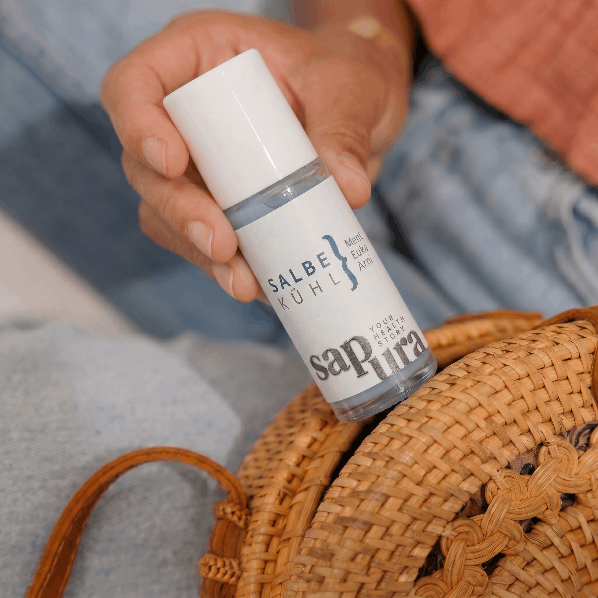 Hand hält sapura health® Kühlgel Roll-On über einer Korbtasche.
