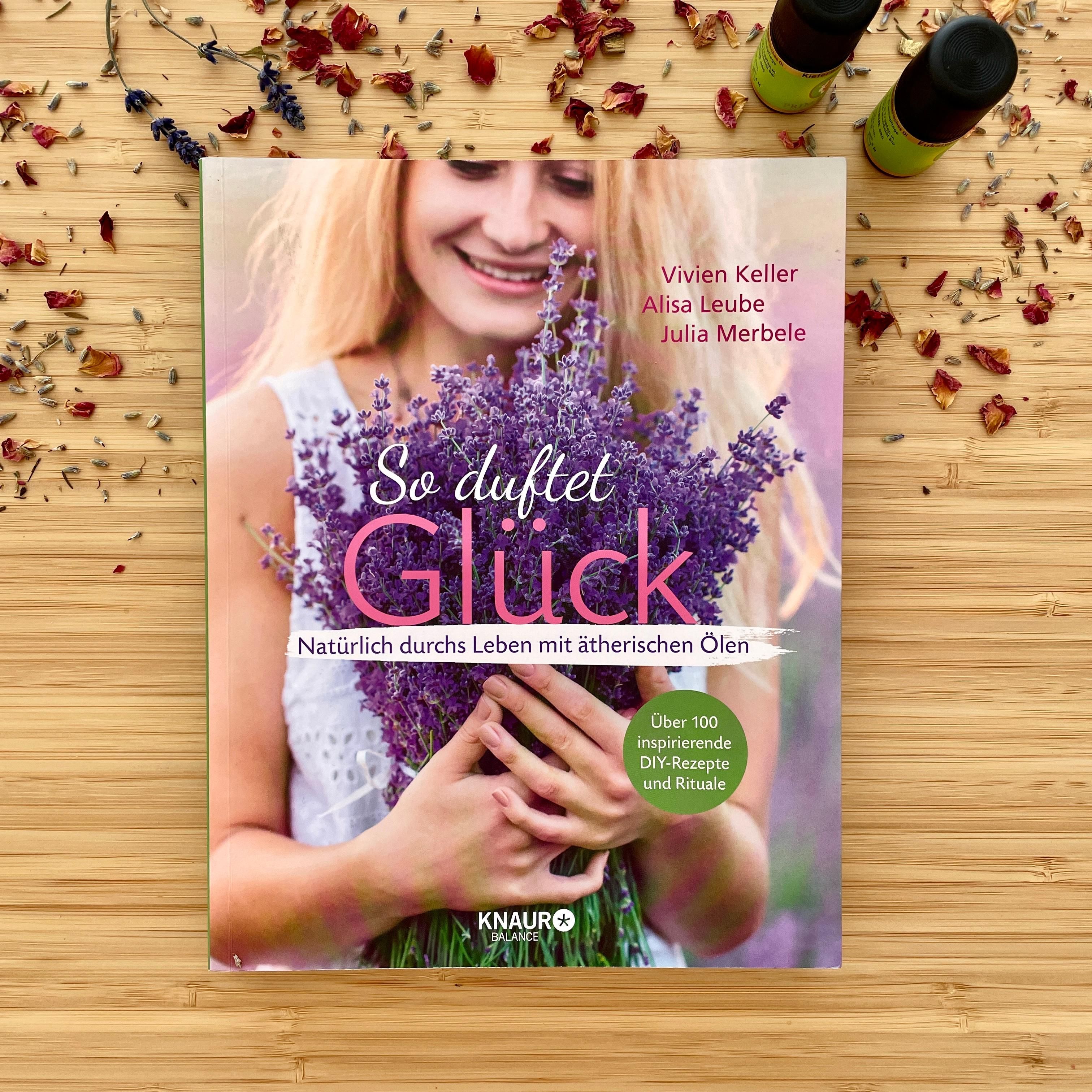 Buch "So duftet Glück" auf Holzuntergrund. Lavendel und ätherische Öle umgeben das Buch. Autoren: Vivien Keller, Alisa Leube, Julia Merbele.