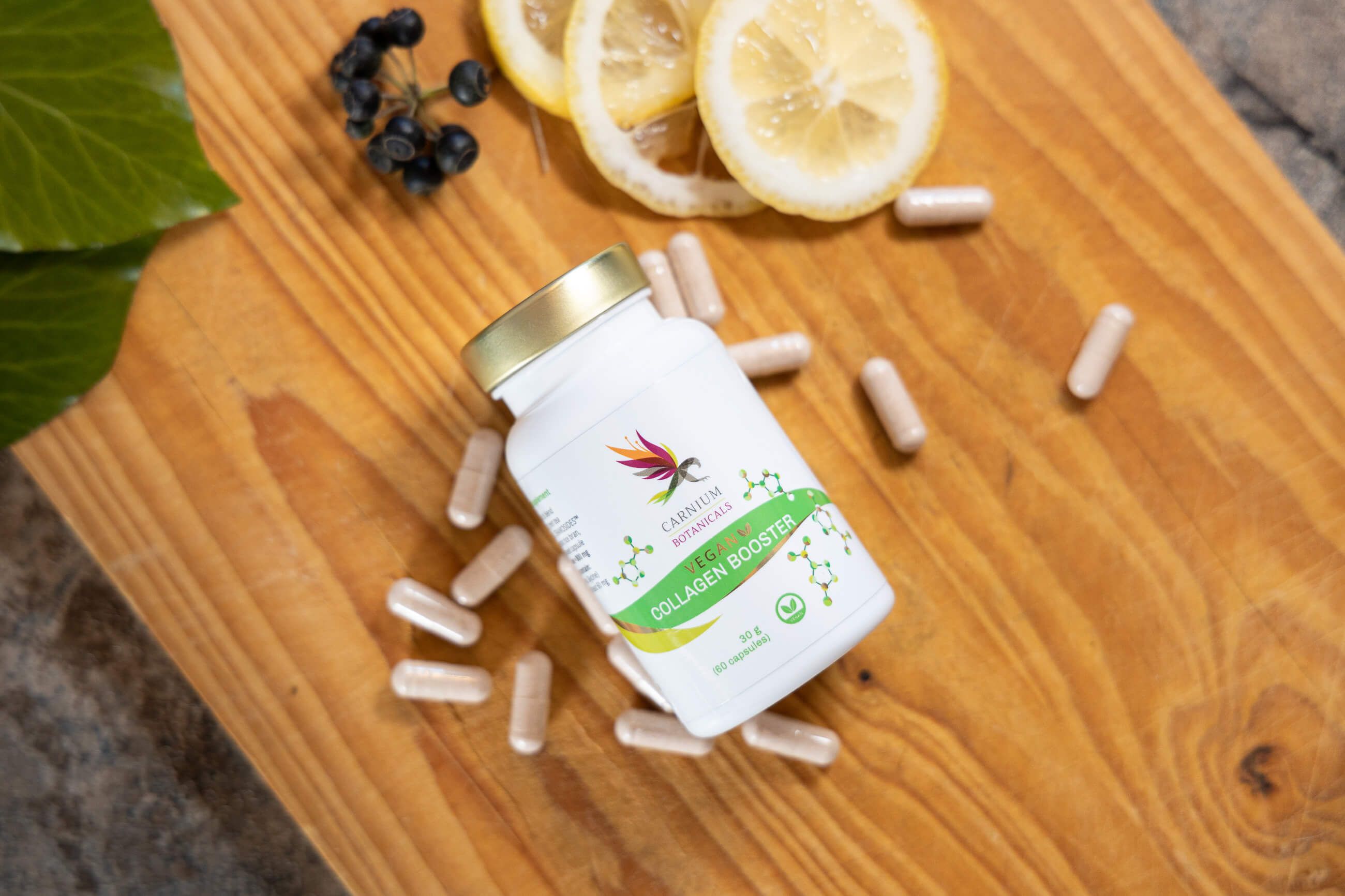 Flasche mit Kapseln auf Holzbrett. Aufschrift: Carnium Botanicals Vegan Collagen Booster. Zitronenscheiben und Beeren.