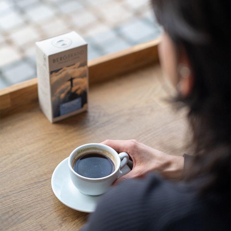 Person hält eine Kaffeetasse und eine Untertasse. Daneben eine Packung BERGBRAND Gipfelkreuz Specialty Coffee.