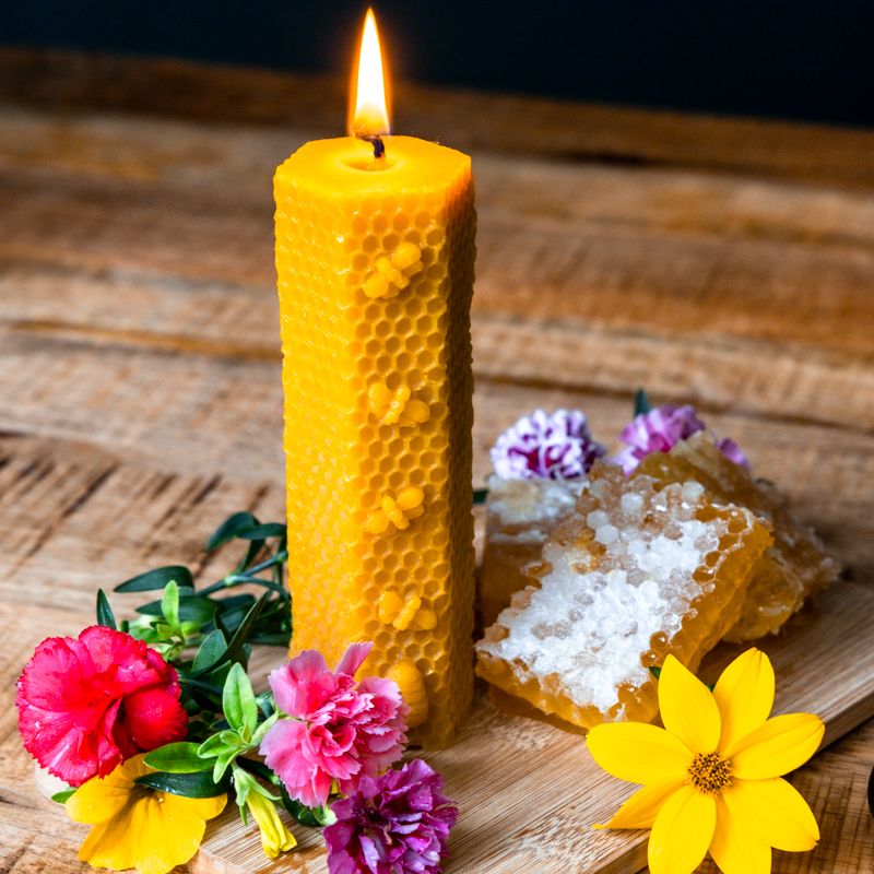 Gelbe Wachskerze mit Bienenwabenmuster, brennend. Neben Honigwaben und Blumen auf Holzbrett.