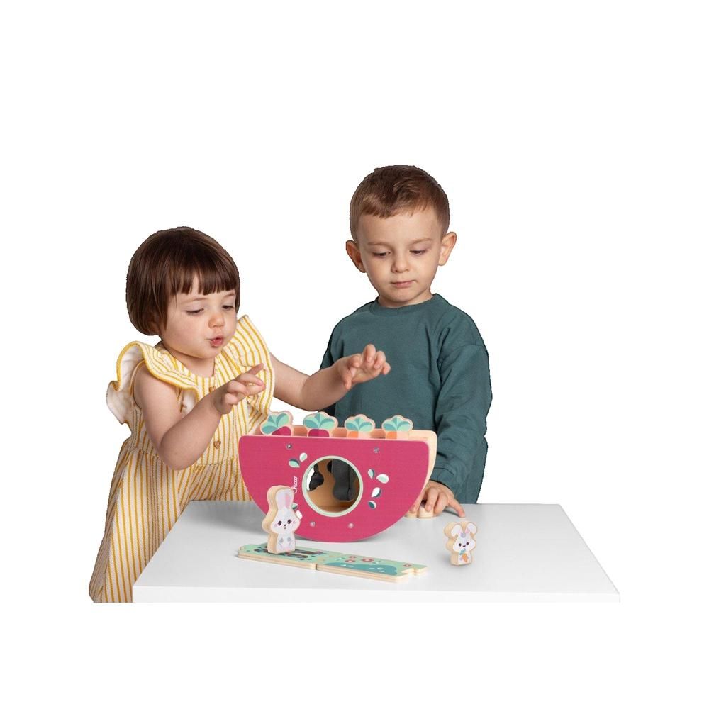 Rosa Holzwippe mit bunten Elementen und Hasenfiguren. Zwei Kinder spielen damit. Auf einer weißen Tischplatte.