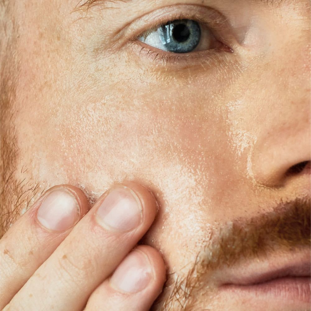 Nahaufnahme eines Mannes, der sein Gesicht berührt. Haut wirkt feucht und glänzend. Bart und Augenpartie sind sichtbar.