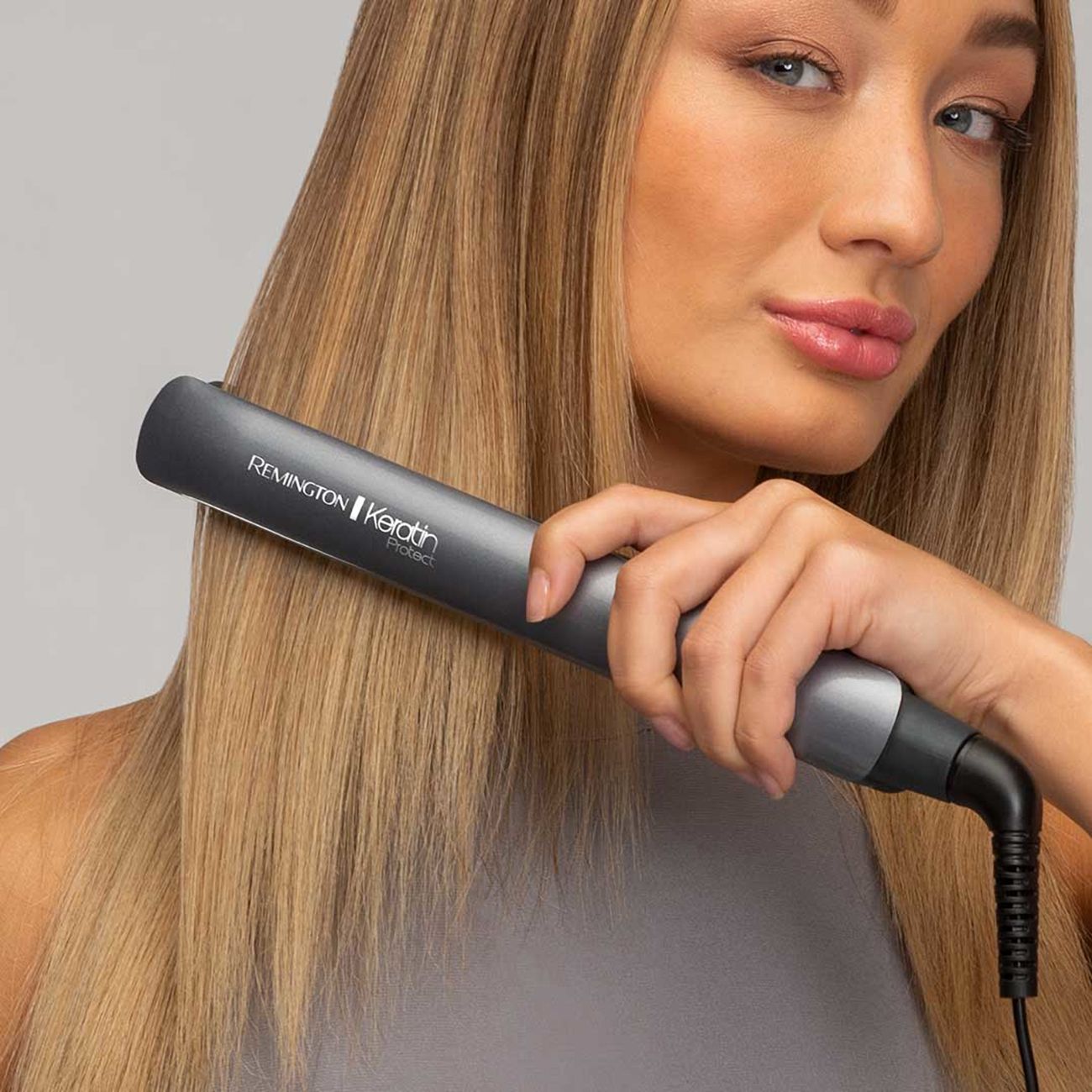 Frau glättet ihr Haar mit einem grauen Glätteisen. Haare sind blond.