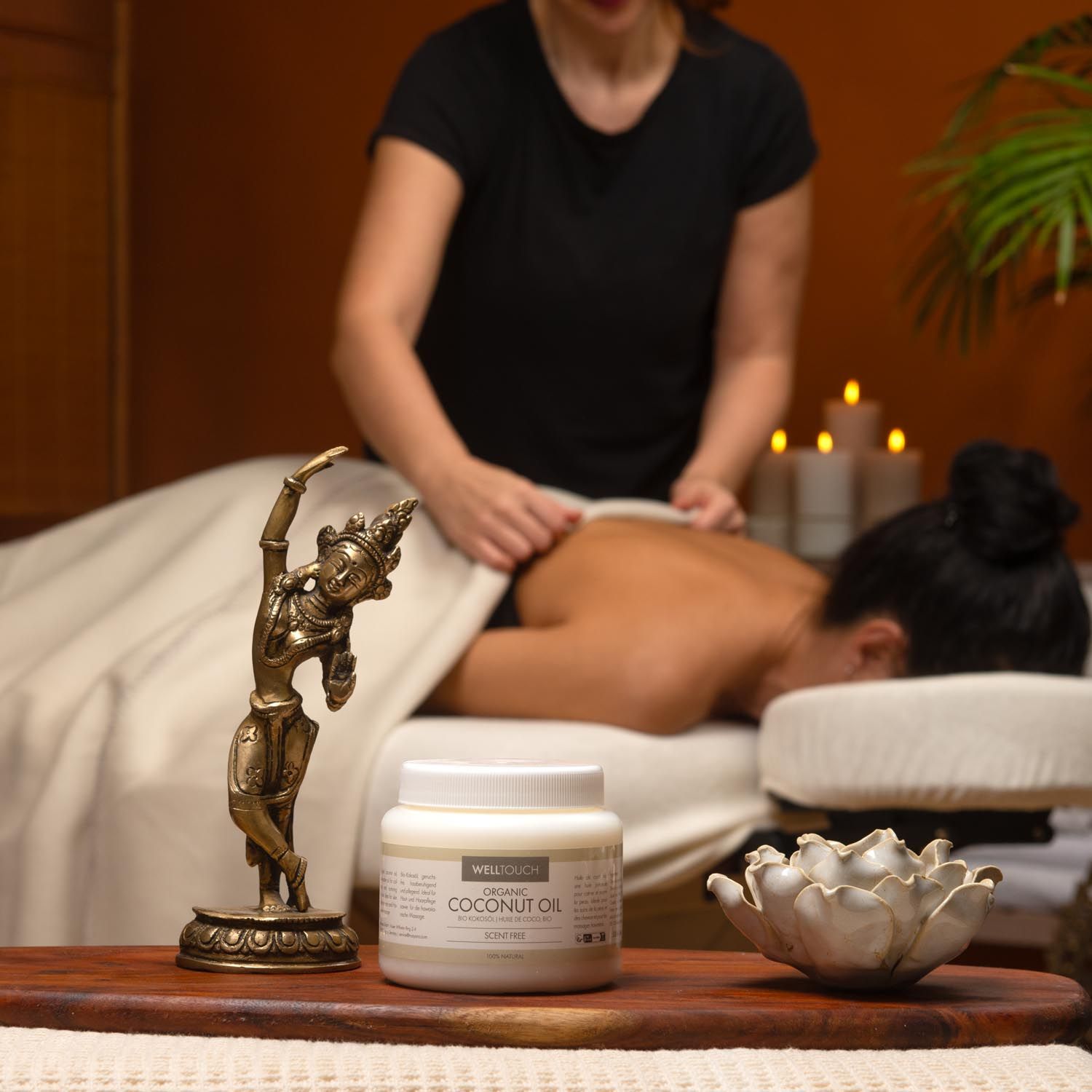 Weißes Glas mit Aufschrift COCONUT OIL. Person erhält Massage. Bronze-Figur und Lotusblüte auf Holzbrett.