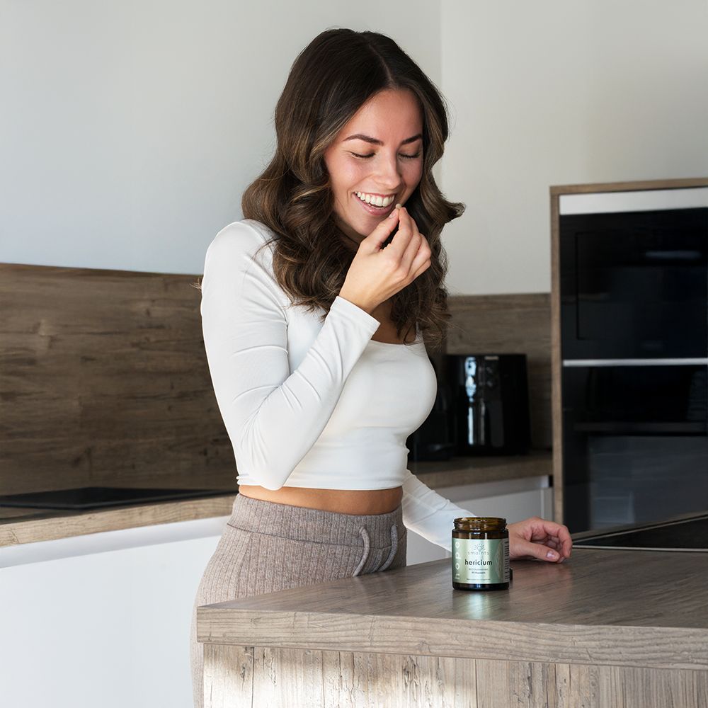 Frau hält eine Kapsel in der Hand, lächelt. Auf dem Tisch steht eine Dose smaints® Hericium.