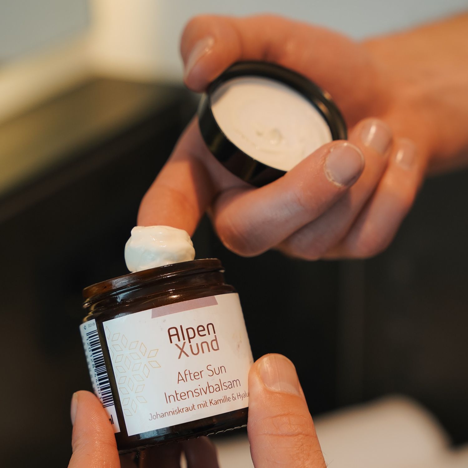 Braune Dose AlpenXund After Sun Intensivbalsam, Deckel geöffnet, Produkt entnommen. Hände halten Dose und Deckel.