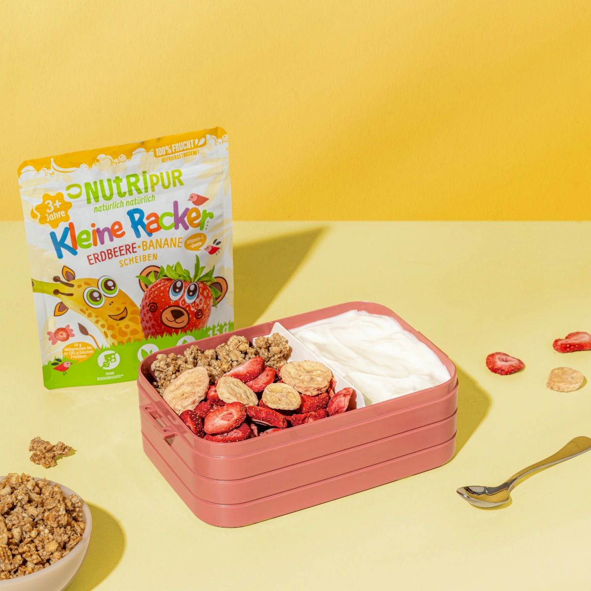 Verpackung mit gefriergetrockneten Früchten und Bento-Box. Aufschrift: NutriPur, Kleine Racker Erdbeere-Banane. Box mit Joghurt und Früchten.