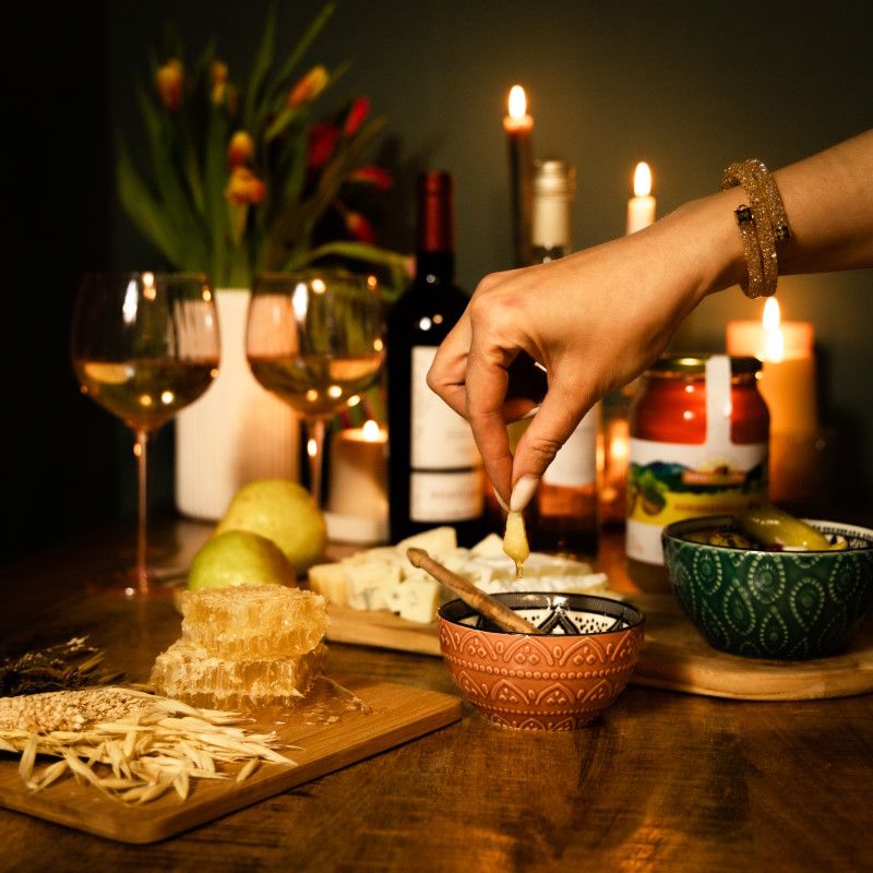 Hand taucht Honig aus einer Schale. Honigwabe, Käse, Wein und Kerzen auf einem Tisch.
