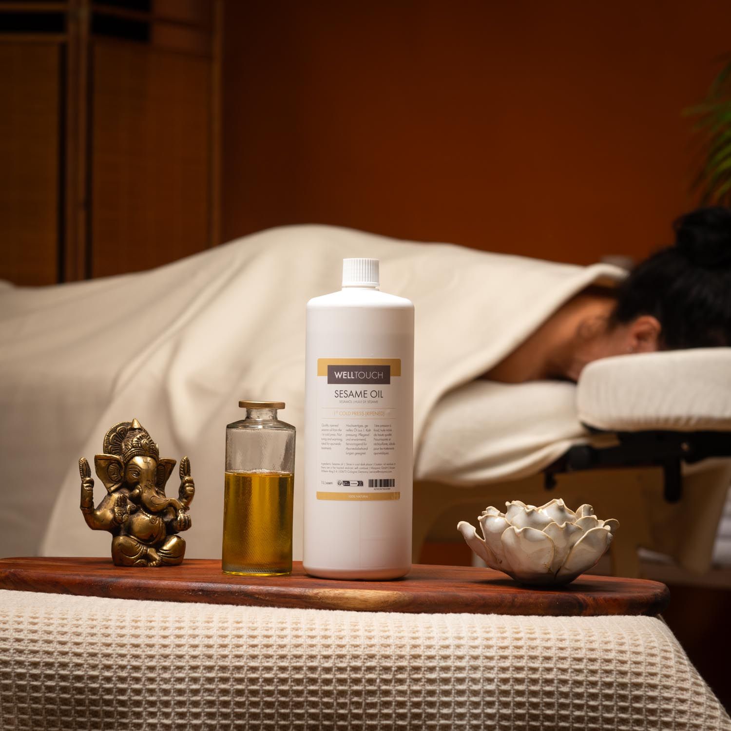 Welltouch Sesamöl-Flasche, Glasflasche, goldfarbenes Öl, Holztablett, Buddha-Figur, Lotusblüte, Massage-Liege.