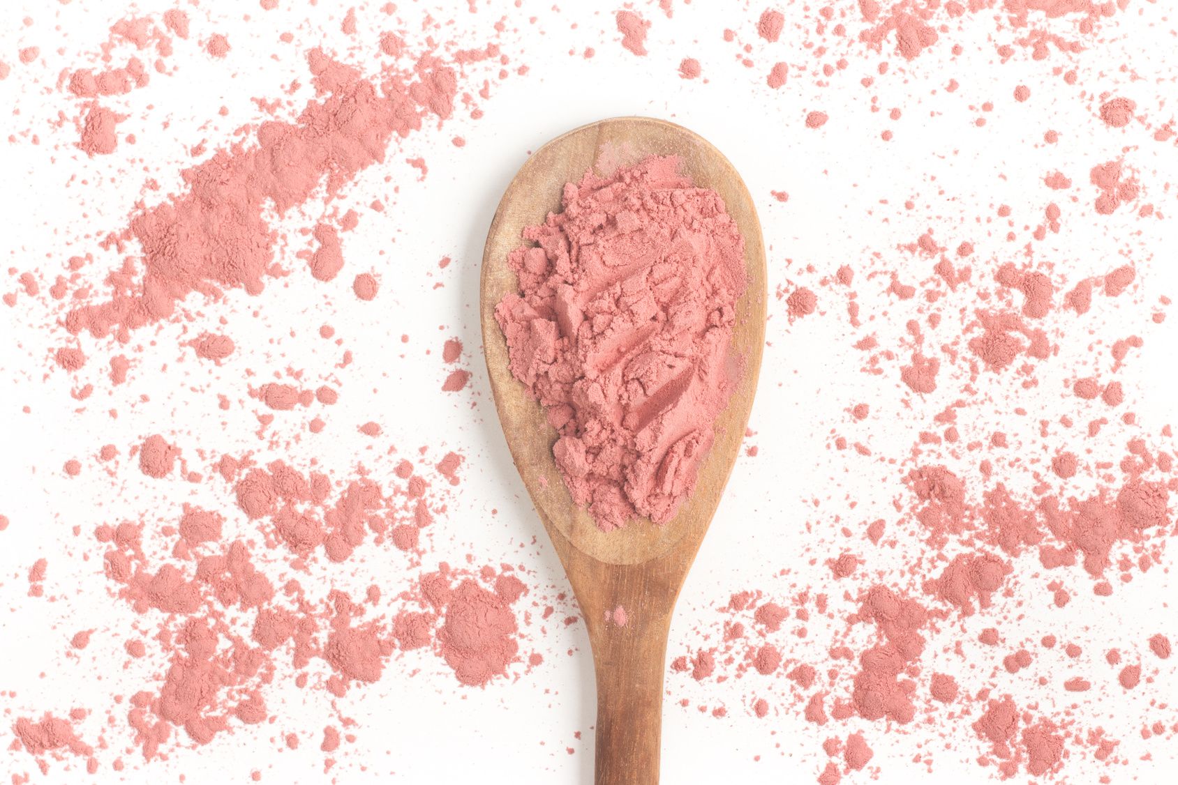 Holzlöffel mit rosa Pulver, umgeben von verstreutem Pulver auf weißem Hintergrund.