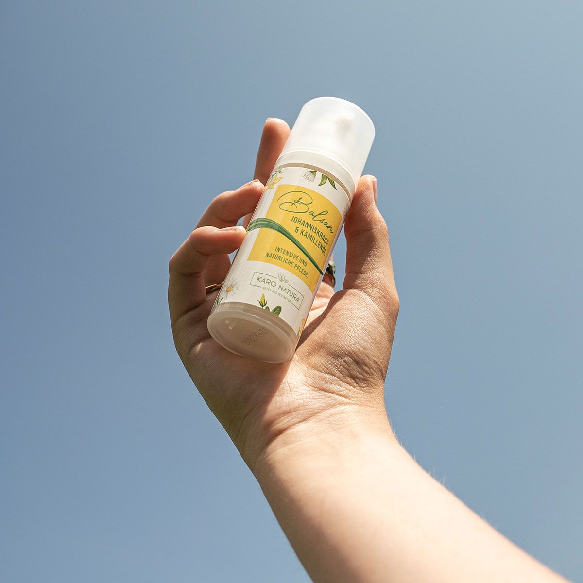 Hand hält eine weiße Flasche mit gelbem Etikett. Aufschrift: Balsam Johanniskraut- & Kamillenöl. Marke: Karo Natura.