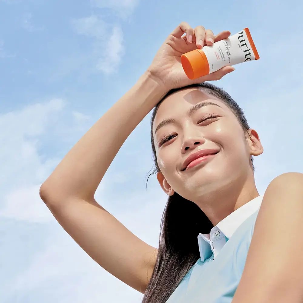 Frau hält eine Tube Sonnencreme über ihrem Kopf. Blauer Himmel im Hintergrund. Die Frau lächelt.