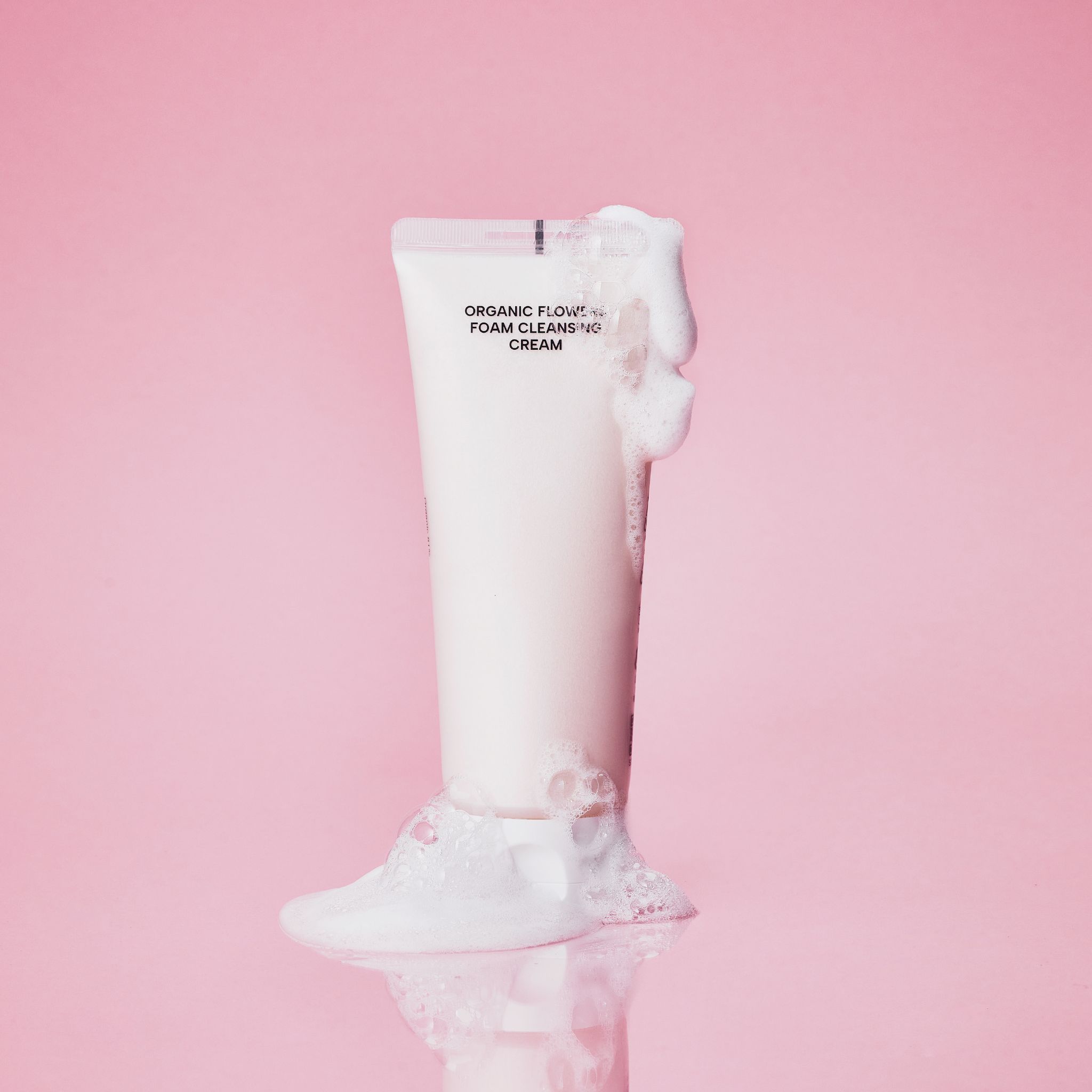 Tubenbehälter mit Schaum, Schriftzug "ORGANIC FLOWERS FOAM CLEANSING CREAM". Rosa Hintergrund.