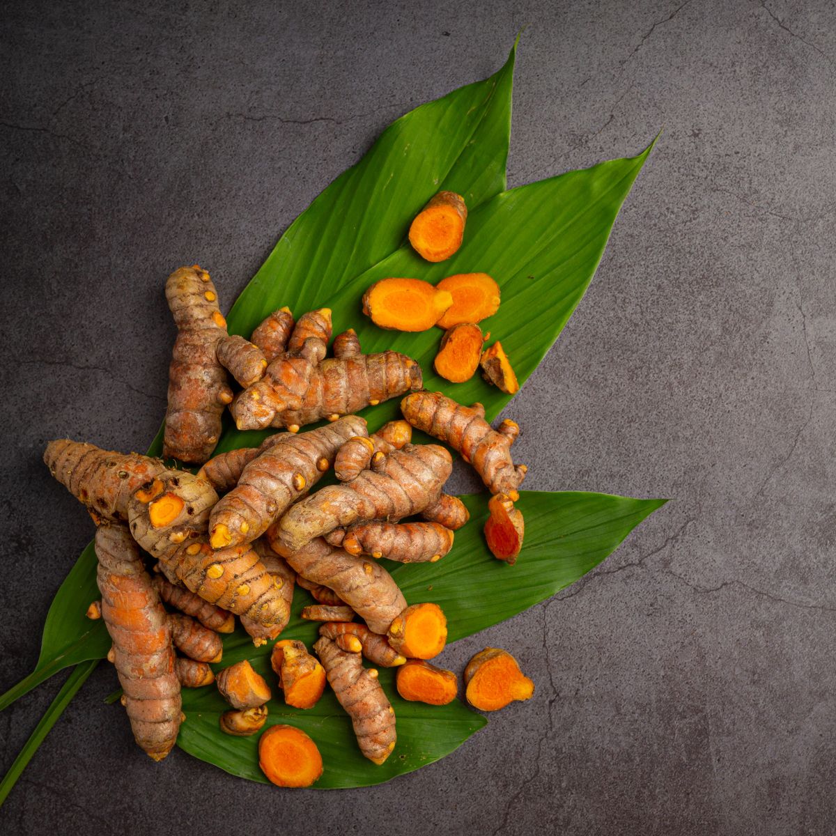 Kurkuma-Wurzeln und -Scheiben auf grünen Blättern, auf dunklem Untergrund. Ansicht von oben.