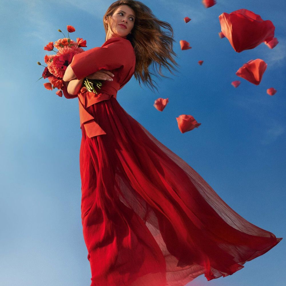 Frau in rotem Kleid fliegt mit einem Blumenstrauß, umgeben von roten Blütenblättern.