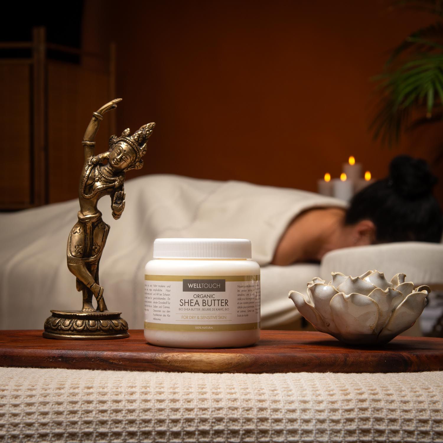 Weißes Glas mit Welltouch Sheabutter. Auf Holztablett, neben goldener Figur und Lotusblüte. Im Hintergrund Person auf Massageliege.
